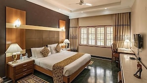 A hotel room with a large bed, bedside tables, a desk, a chair, a ceiling fan, and a window with curtains. - Eagleton The Golf Resort (Ramanagara)