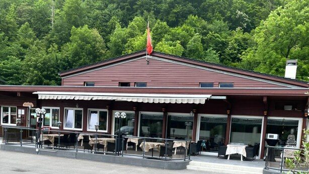 A building with a red roof and a flagpole, surrounded by a parking lot and greenery. - The River Village (Interlaken)