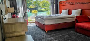A hotel room with a bed, a red sofa, a wooden dresser, and a view of a river. - The River Village (Interlaken)