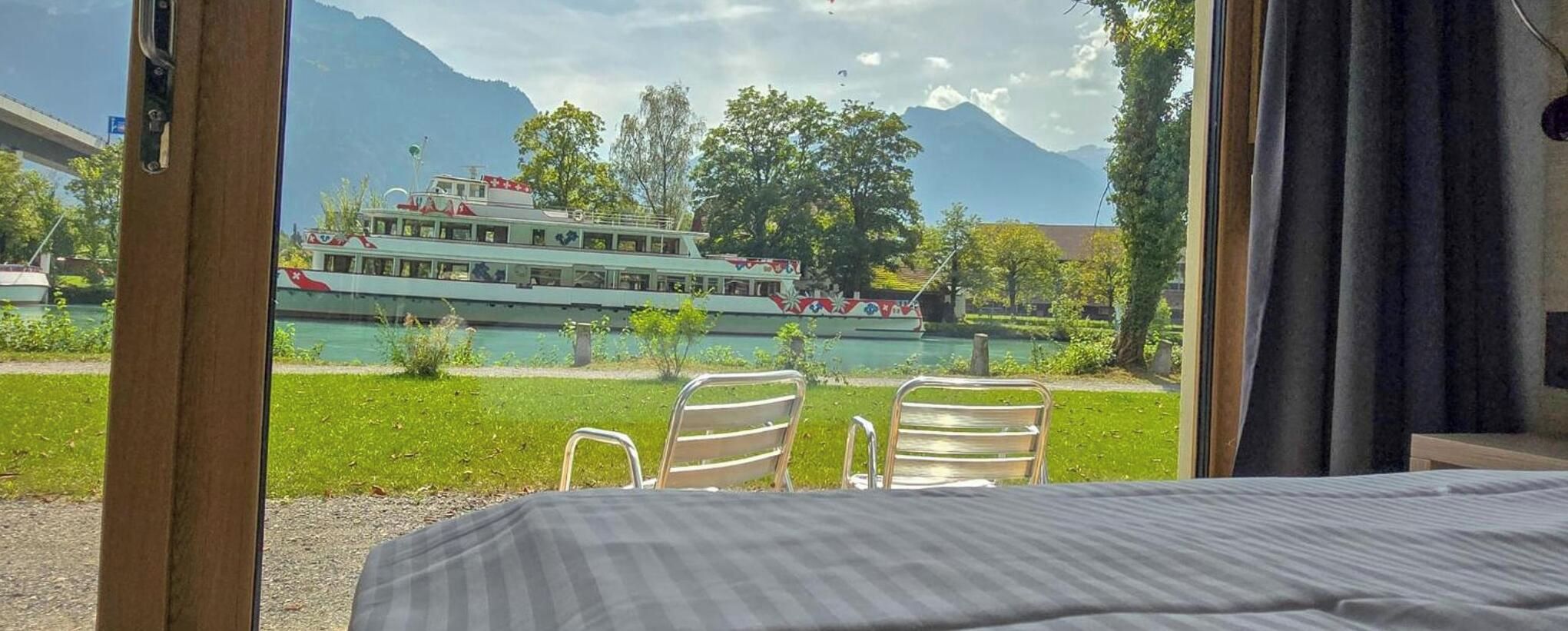 Ein Zimmer mit einem Bett, zwei Stühlen und Blick auf einen See mit einem Boot und Bergen.