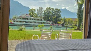 A room with a bed, two chairs, and a view of a lake with a boat and mountains. - The River Village (Interlaken)