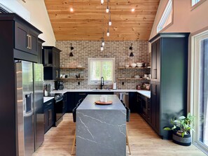 A modern kitchen with a central island, black cabinetry, a brick wall, and pendant lighting. - New Lake Front Aframe Cabin/Kayaks/Arcade/Fishing/Mountain Views/Hot Tub (Ranger)