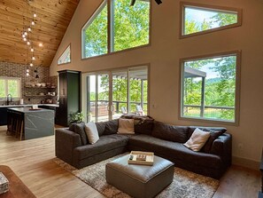 A modern living room with a sectional sofa, a coffee table, and large windows offering views of greenery. - New Lake Front Aframe Cabin/Kayaks/Arcade/Fishing/Mountain Views/Hot Tub (Ranger)