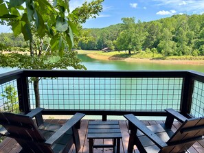 A wooden deck with two chairs and a table overlooking a calm lake, surrounded by lush greenery and a hilly landscape.
