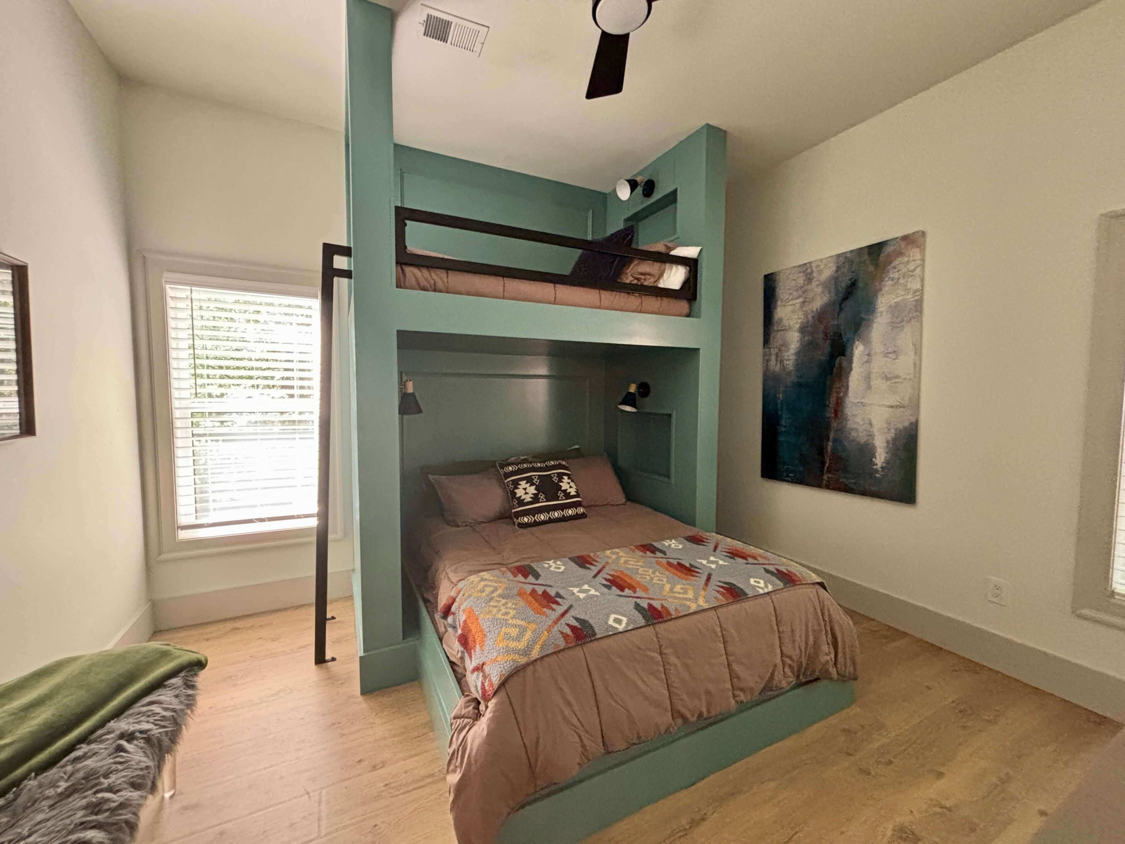 Una habitación con literas, una ventana con persianas, un cuadro en la pared y un ventilador de techo.
