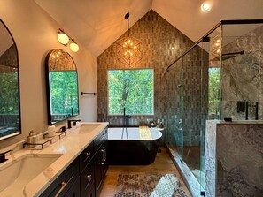 A modern bathroom with a large bathtub, a glass-enclosed shower, and two large windows offering views of a forest.