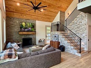 A modern living room with a fireplace, a stone wall, and a wooden ceiling. - New Lake Front Aframe Cabin/Kayaks/Arcade/Fishing/Mountain Views/Hot Tub (Ranger)