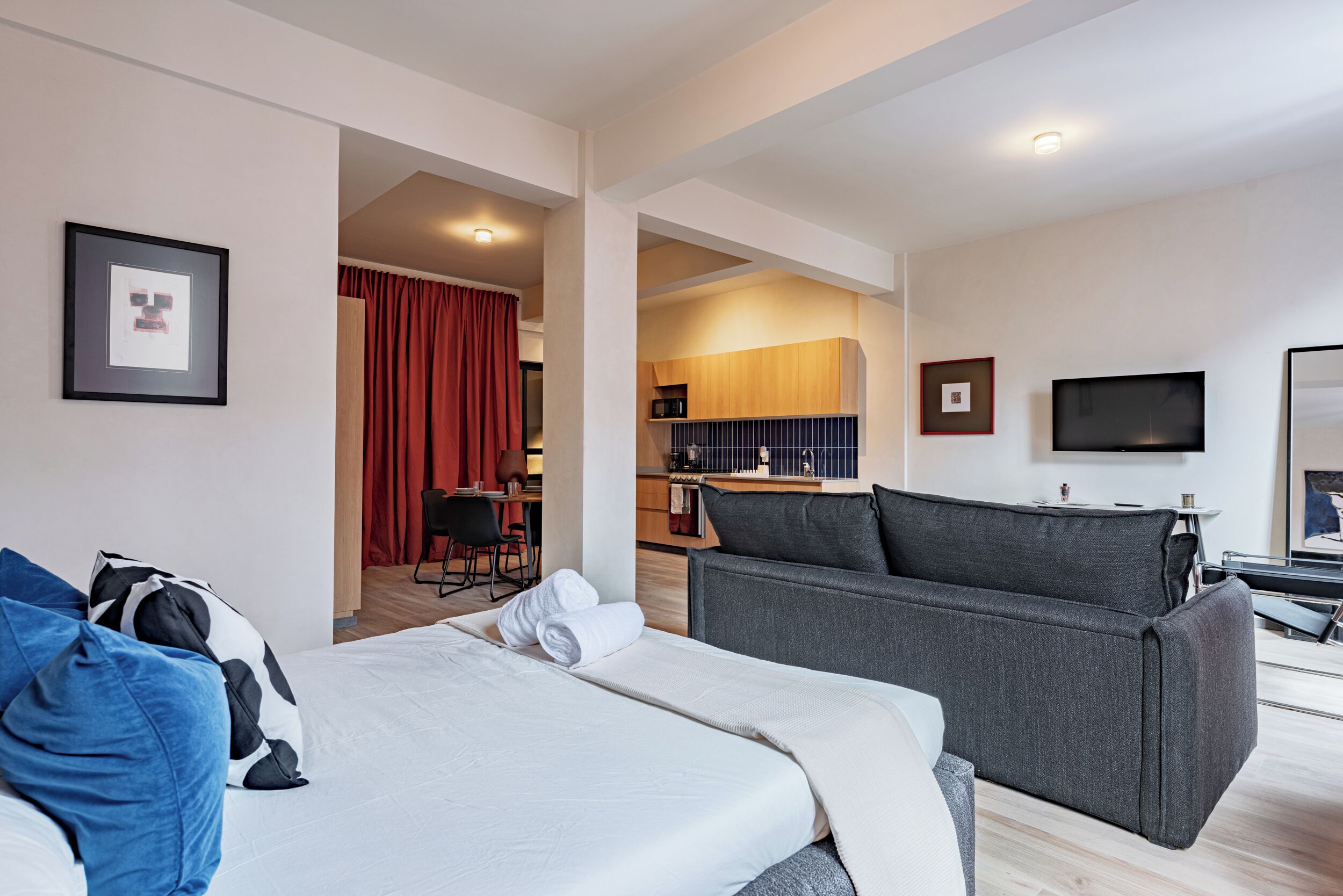 A modern hotel room with a bed, a sofa, a TV, and a kitchenette.