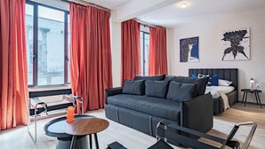 Een moderne woonkamer met een grijze bank, rode gordijnen, een kleine ronde tafel en een bed met een wit hoofdeinde.