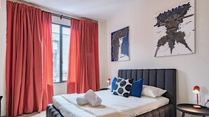 Een moderne hotelkamer met een groot bed, rode gordijnen, een hoofdeinde, een nachtkastje met een lamp en een kunstwerk aan de muur.