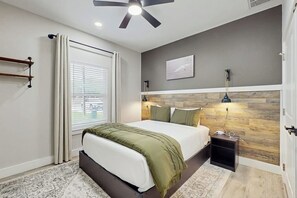 Ein Schlafzimmer mit einem Bett, einem Deckenventilator, einem Nachttisch, einer Lampe, einem Fenster mit Jalousien und einem Wandbild.