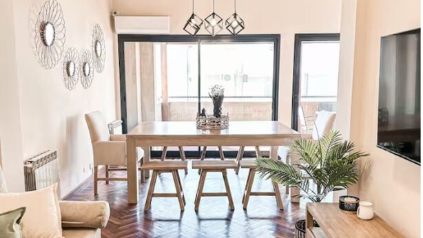 A spacious living area with a wooden herringbone floor, a central dining table with chairs, a sofa, a coffee table, and a TV mounted on the wall. - Lujoso amplio dpto para 8 en Salta con cochera (Salta)