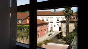 Visão pela janela de um edifício com um pátio, mostrando uma estrutura de telhado vermelho e um prédio branco com várias janelas.