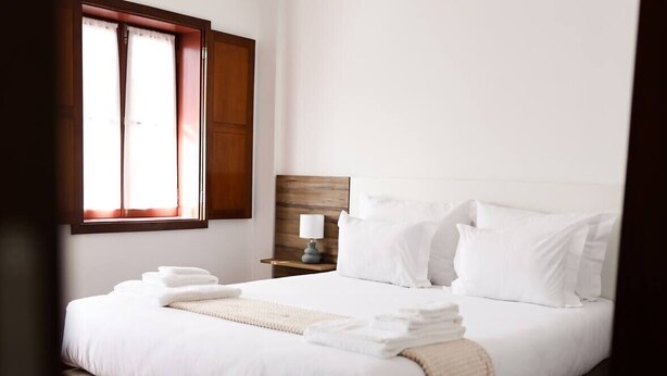 Cama bem arrumada com lençóis brancos e manta bege, mesa de cabeceira de madeira com abajur, e janela com cortinas.