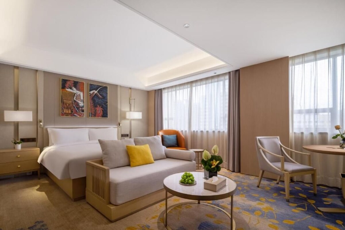 A hotel room with a bed, a sofa, a coffee table, a chair, a lamp, and a painting on the wall.