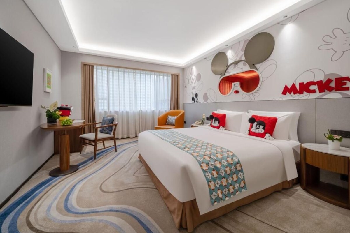 A hotel room with a bed, a desk, a chair, a TV, and Mickey Mouse-themed decor.