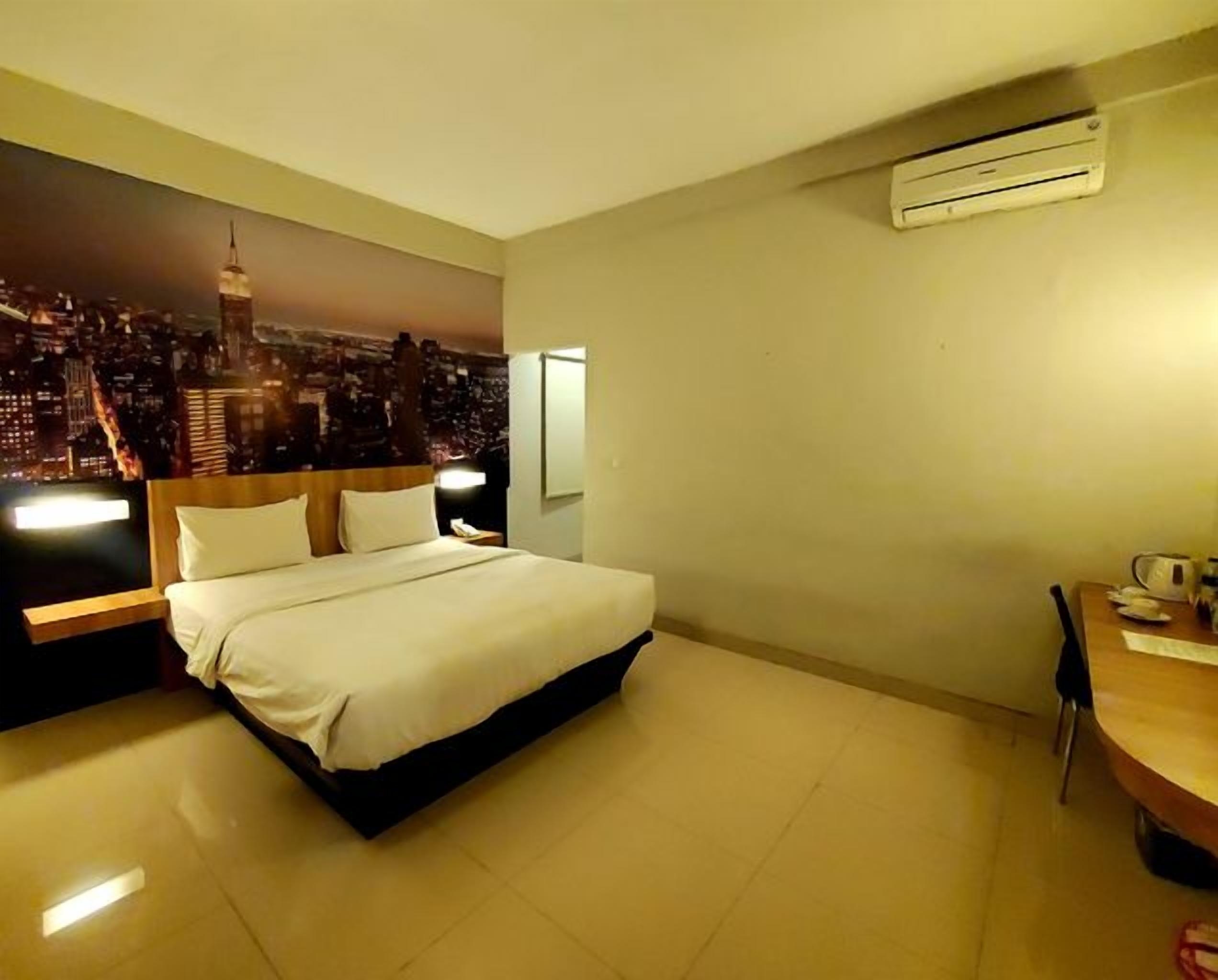 A hotel room with a bed, a nightstand, a desk, and a cityscape mural on the wall.