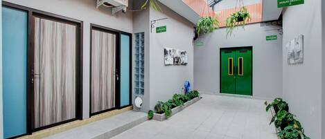 A corridor with a green door, air conditioning units, hanging plants, and a tiled floor.