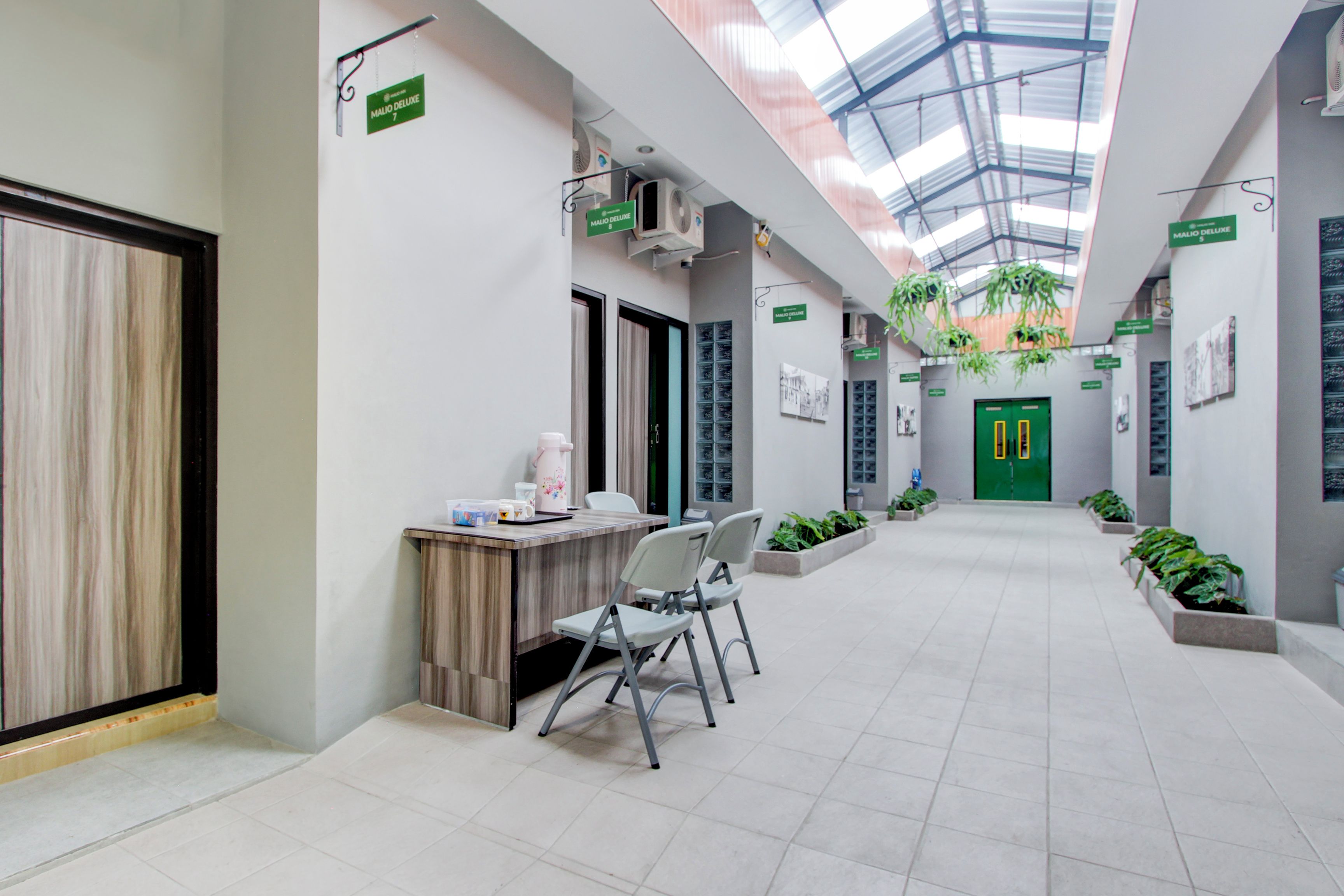 A modern, well-lit hallway with a glass ceiling, green signage, and indoor plants.
