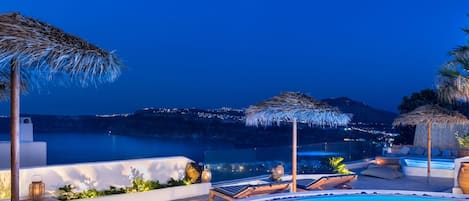 A poolside area with lounge chairs, a pool, and a view of the coastline at night.