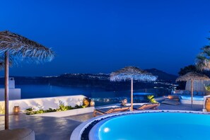 A poolside area with lounge chairs, a pool, and a view of the coastline at night. - Summer Lovers Villa (Santorini)