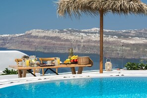 A poolside breakfast setup with a table, fruits, and a view of a mountainous coastline. - Summer Lovers Villa (Santorini)