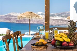 Uma mesa com uma fruteira, um jarro e copos, com vista para uma piscina e o mar.