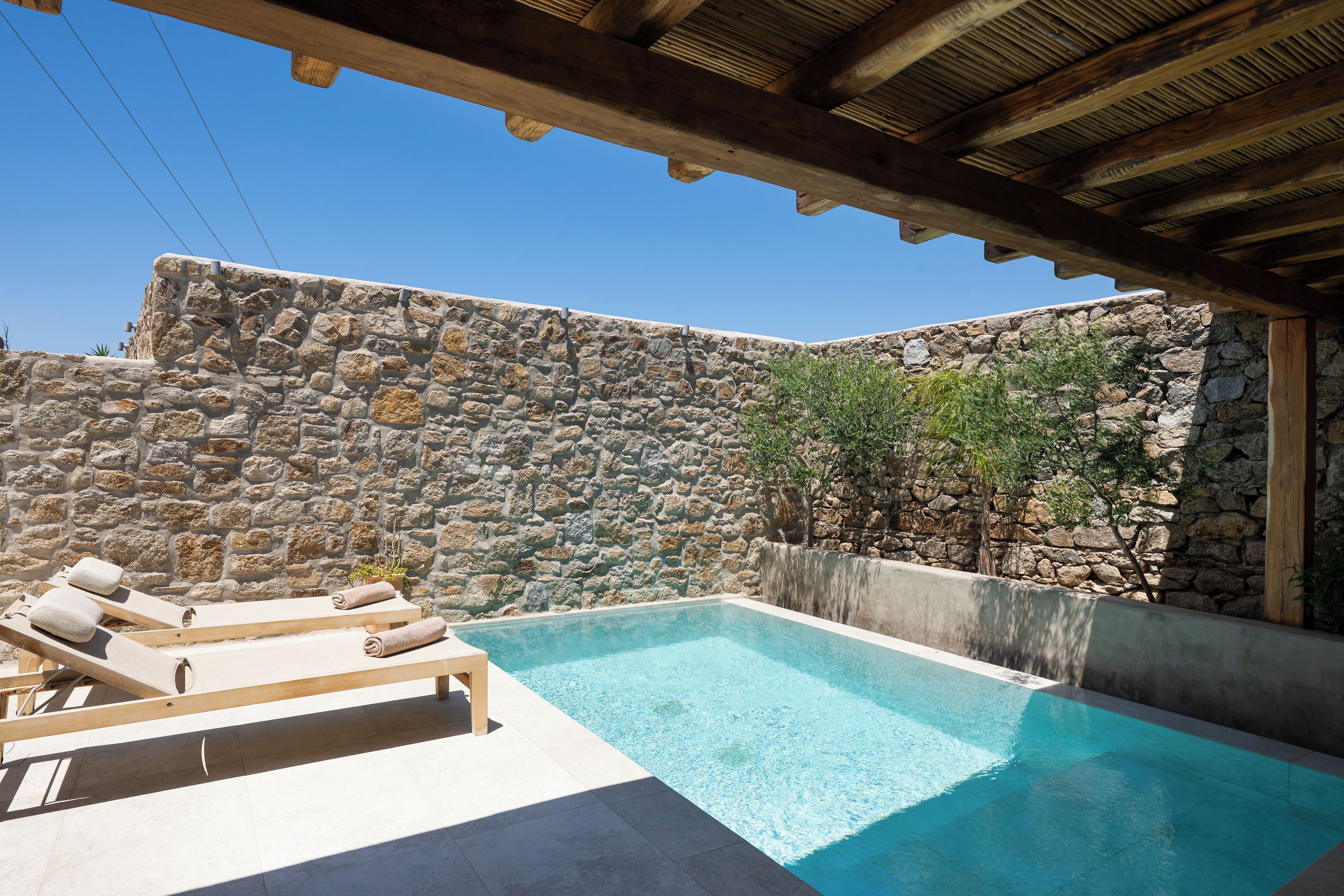Una piscina con sillones de descanso, un muro de piedra y una pérgola de madera.