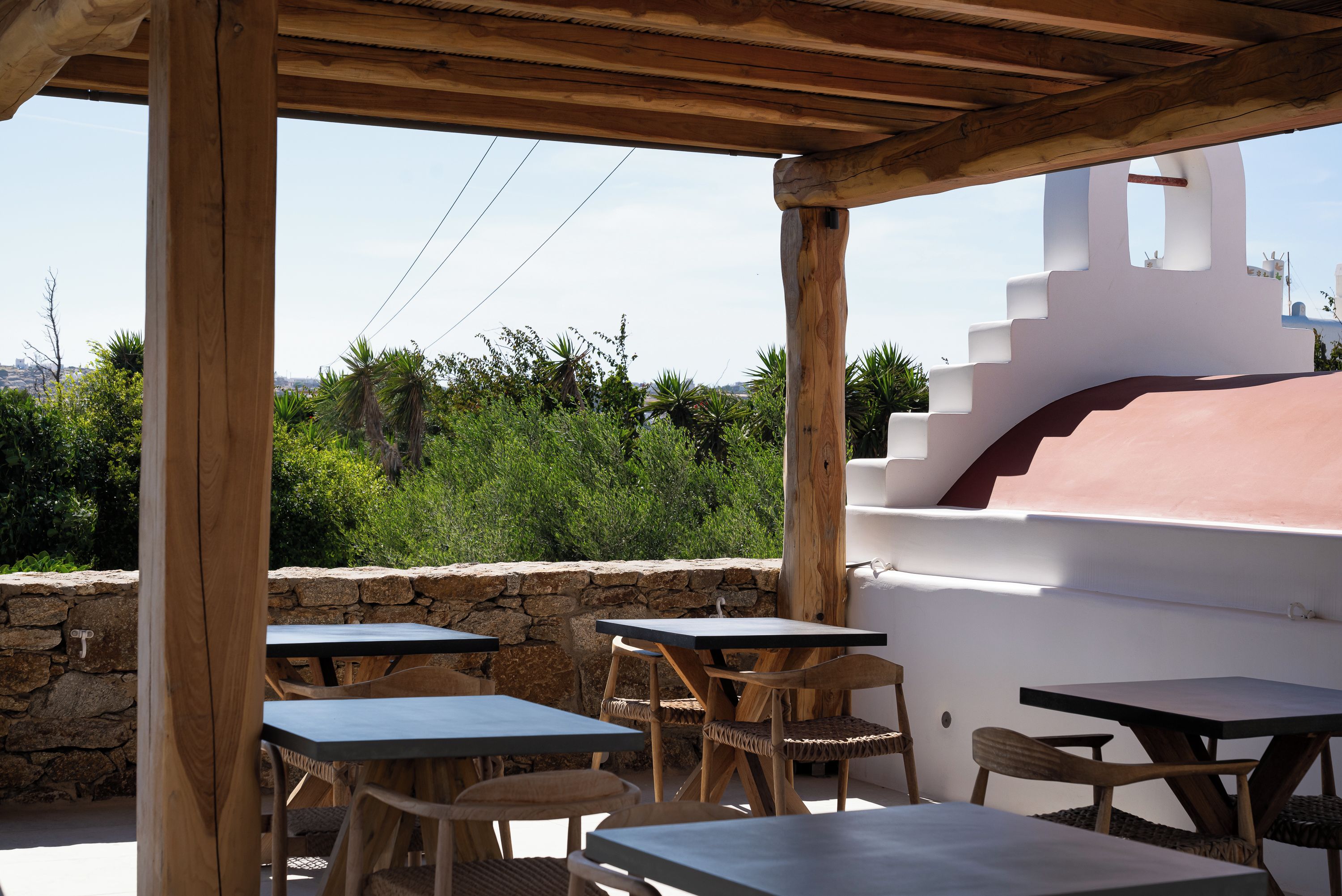 An outdoor patio with wooden tables and chairs, a stone wall, and a view of greenery and a building in the background.