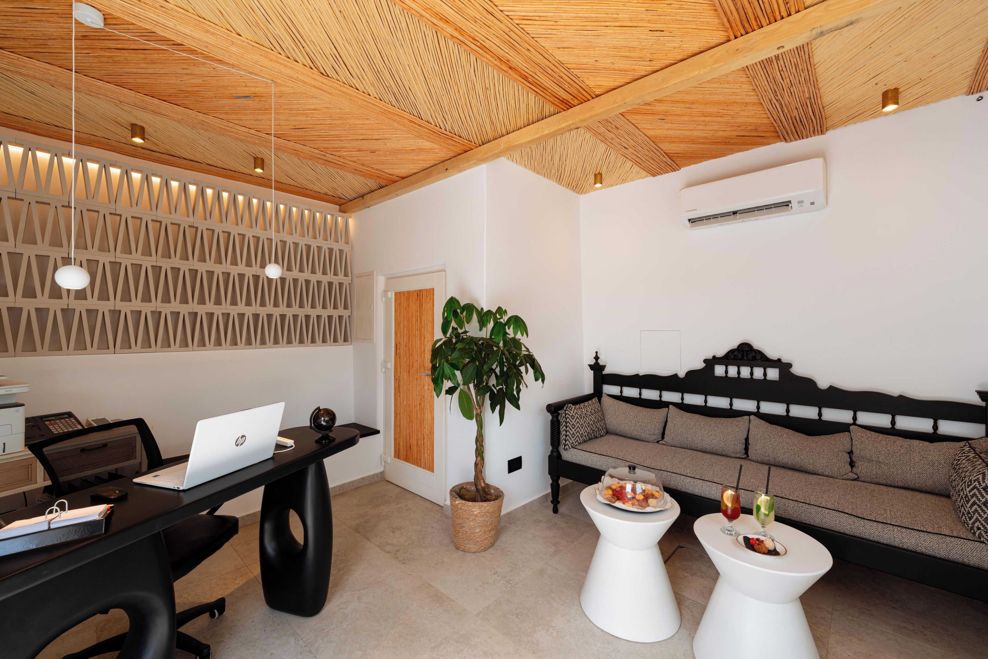 A modern room with a wooden ceiling, a desk with a laptop, a potted plant, a sofa, and two white side tables with drinks.
