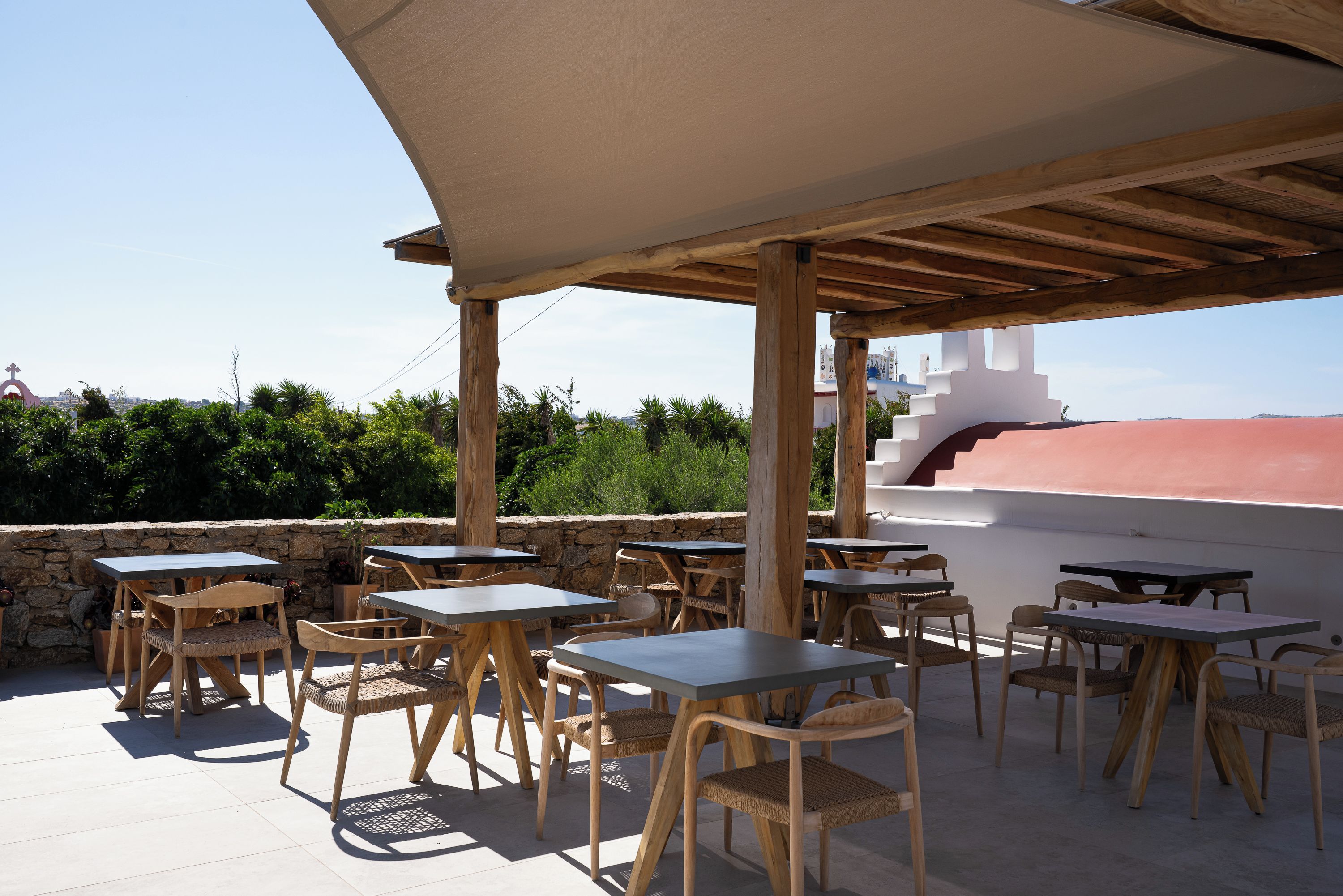 An outdoor patio with wooden tables and chairs, a stone wall, and a view of greenery and buildings in the background.