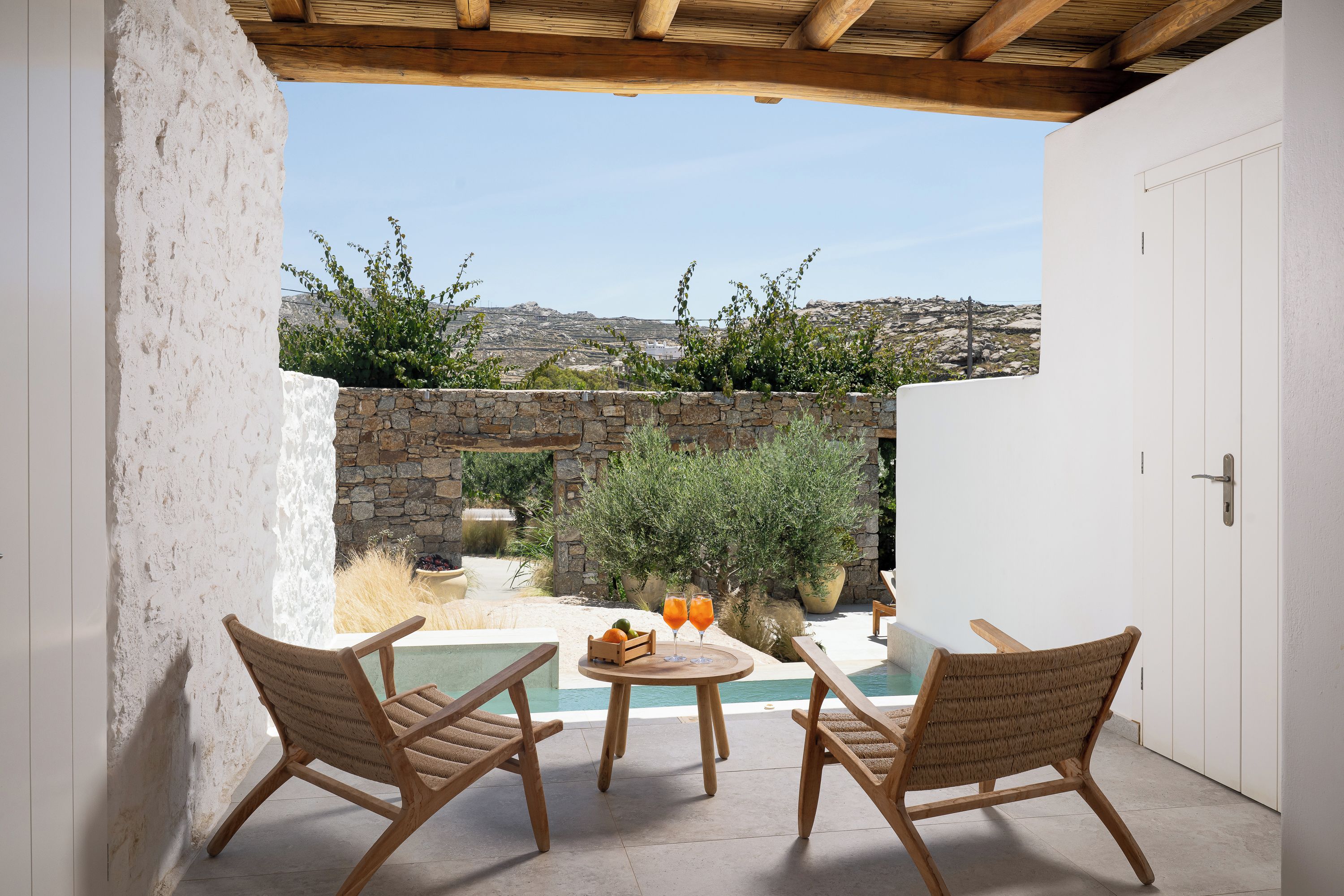 A patio with a pool, two wicker chairs, a table with drinks, and a stone wall.