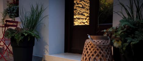 Entrada de hotel con felpudo, plantas en macetas, silla de mimbre y pared texturizada.
