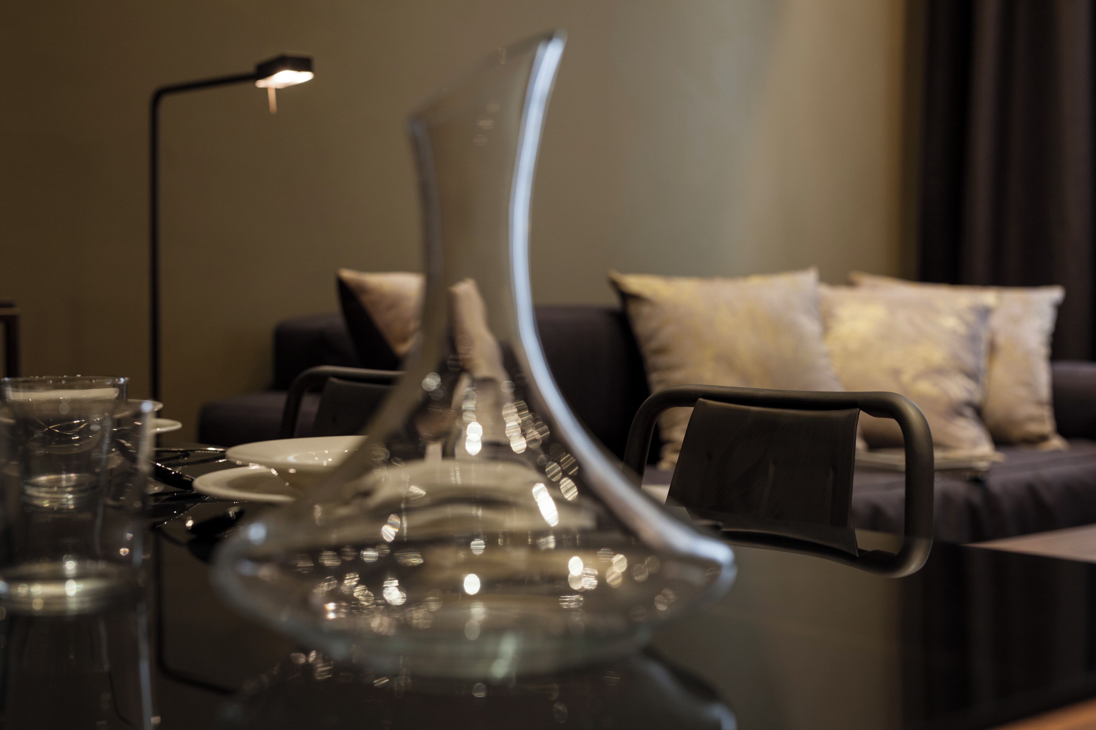 A dining table with a clear glass vase, a black lamp, and a sofa with cushions in the background.