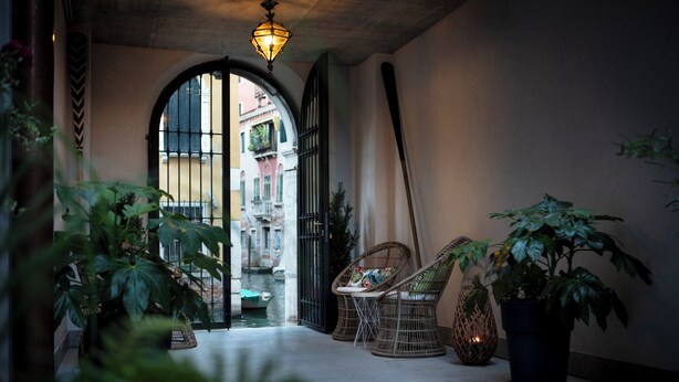 A corridor with wicker chairs, potted plants, and a view of a canal and buildings. - Aqua Apartments (Venice)