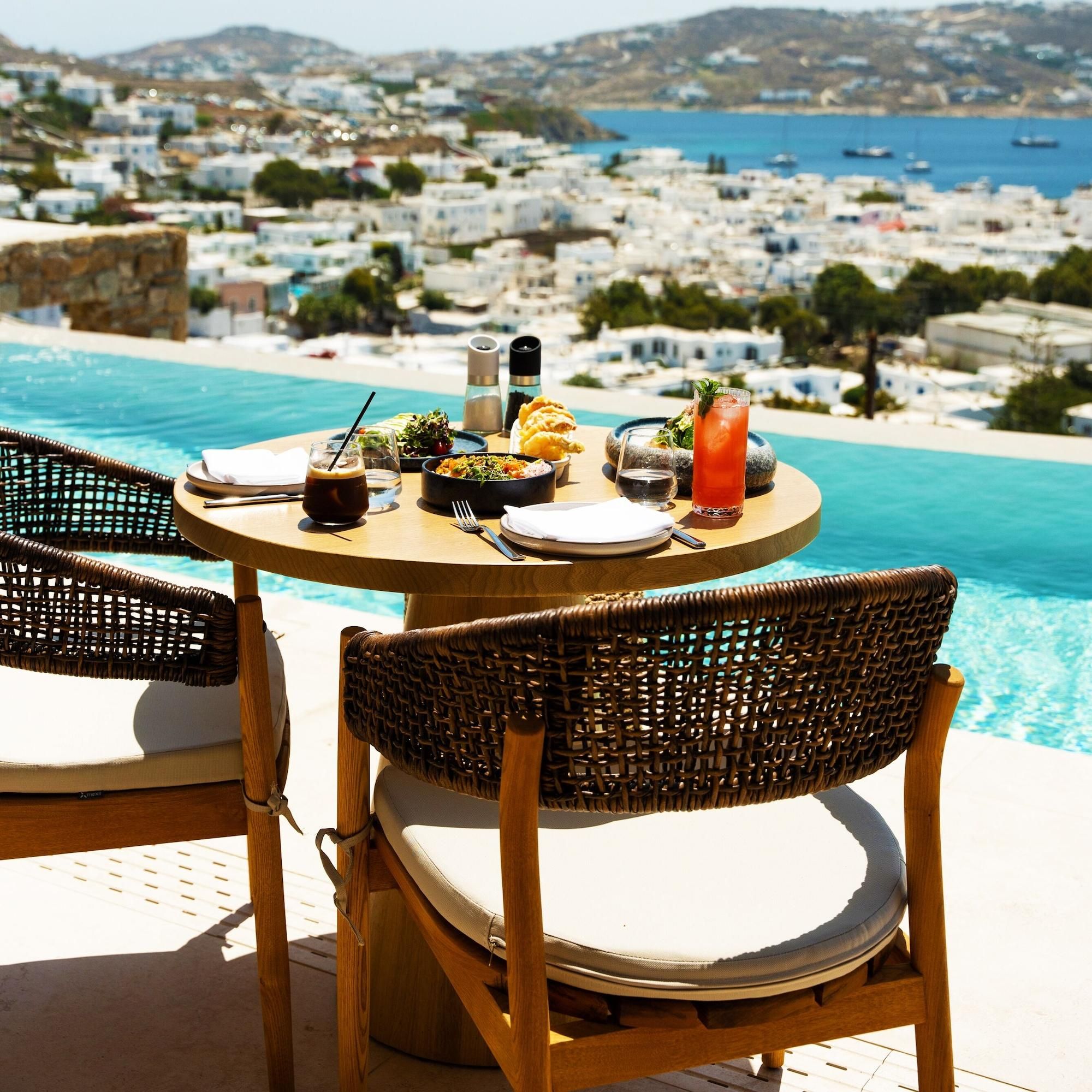 A dining table with a view of a coastal town, two wicker chairs, and a poolside setting.