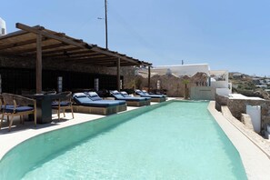 A poolside area with lounge chairs and a pergola, a stone wall, and a view of the landscape. - Numi Boutique Hotel Mykonos (Mykonos)
