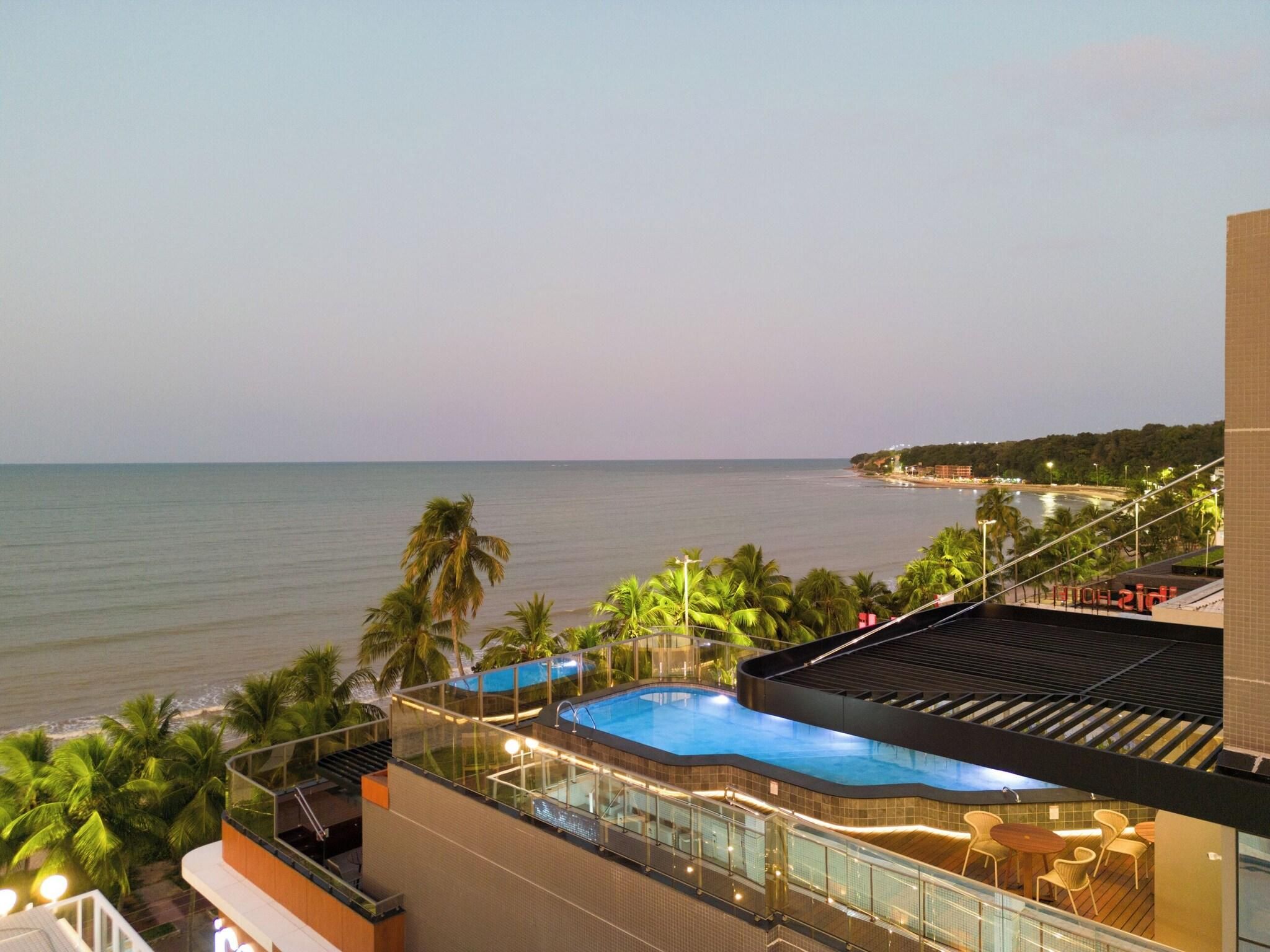 Piscina no terraço com vista para o mar, palmeiras e um edifício com fachada em azulejos.