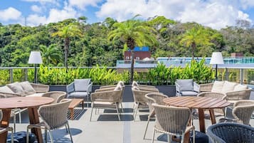 Pátio externo com móveis de vime e mesas, com vista para uma paisagem verdejante e um edifício com telhado azul.