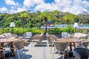 An outdoor patio with wicker furniture and tables, overlooking a lush green landscape and a building with a blue roof. - Icon Hotel By Welkom (Pitimbu)