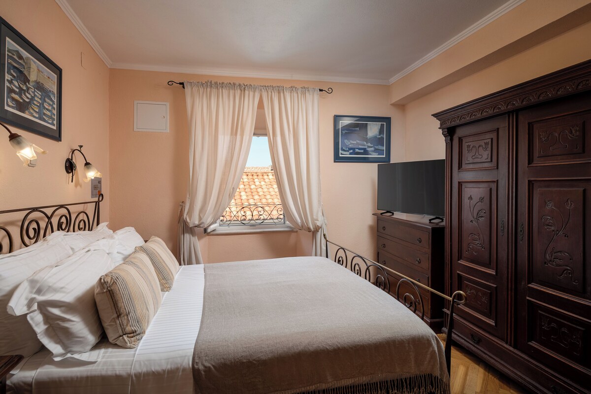 A bedroom with a bed, a dresser, a TV, a window with curtains, and wall-mounted lights.