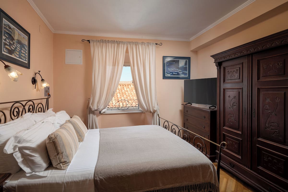 Ein Schlafzimmer mit Bett, Kommode, Fernseher, Fenster mit Vorhängen und Wandleuchten.