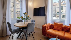 Ein Zimmer mit Esstisch, Stühlen, einer Couch, einem Fernseher und großen Fenstern mit Vorhängen.