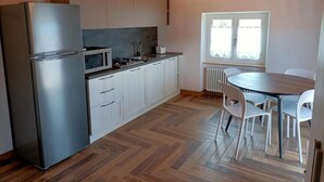 Cocina compacta con refrigerador, microondas y estufa, piso de madera, y zona de comedor con mesa y sillas.
