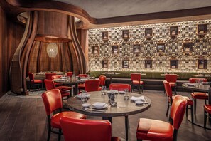 A restaurant with round tables, red chairs, a wooden wall with framed pictures, and a green cushioned seating area. - Nobu New Orleans (New Orleans)