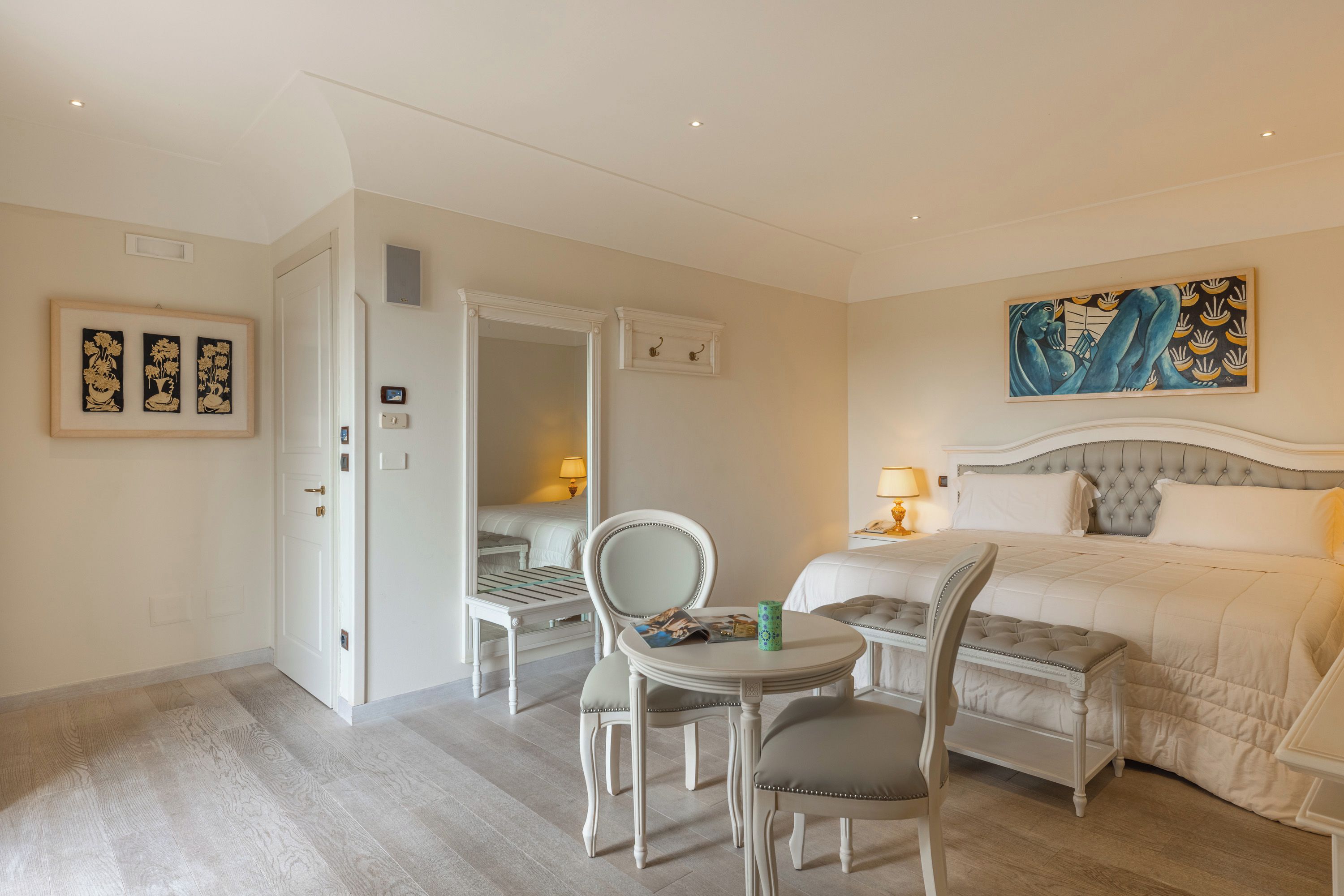 Une chambre d’hôtel comprenant un lit, une table à manger, deux chaises, une petite table avec une lampe et des œuvres d’art accrochées aux murs.