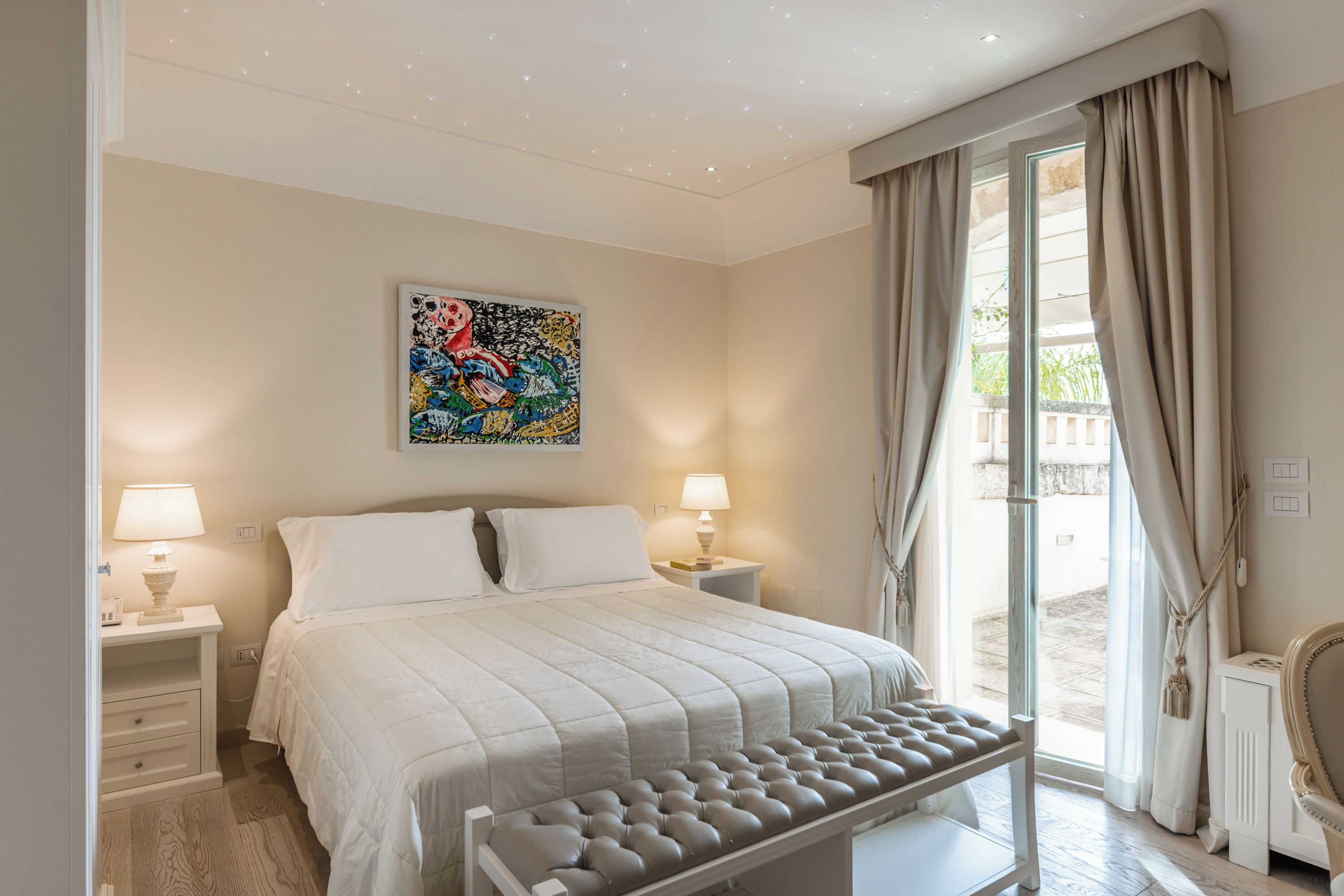 A bedroom with a bed, bedside lamps, a window with curtains, and a wall-mounted artwork.