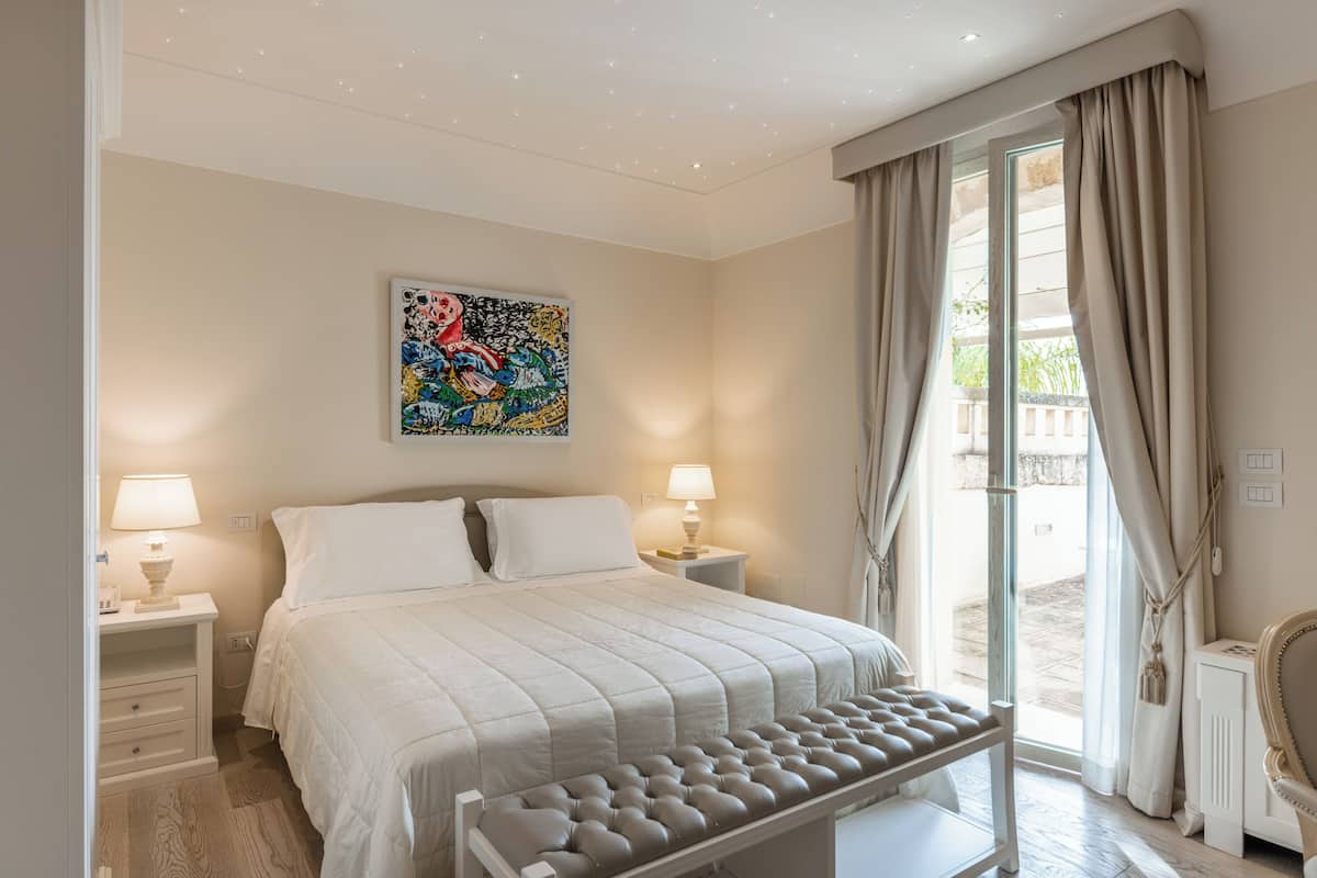 A bedroom with a bed, bedside lamps, a window with curtains, and a wall-mounted artwork.