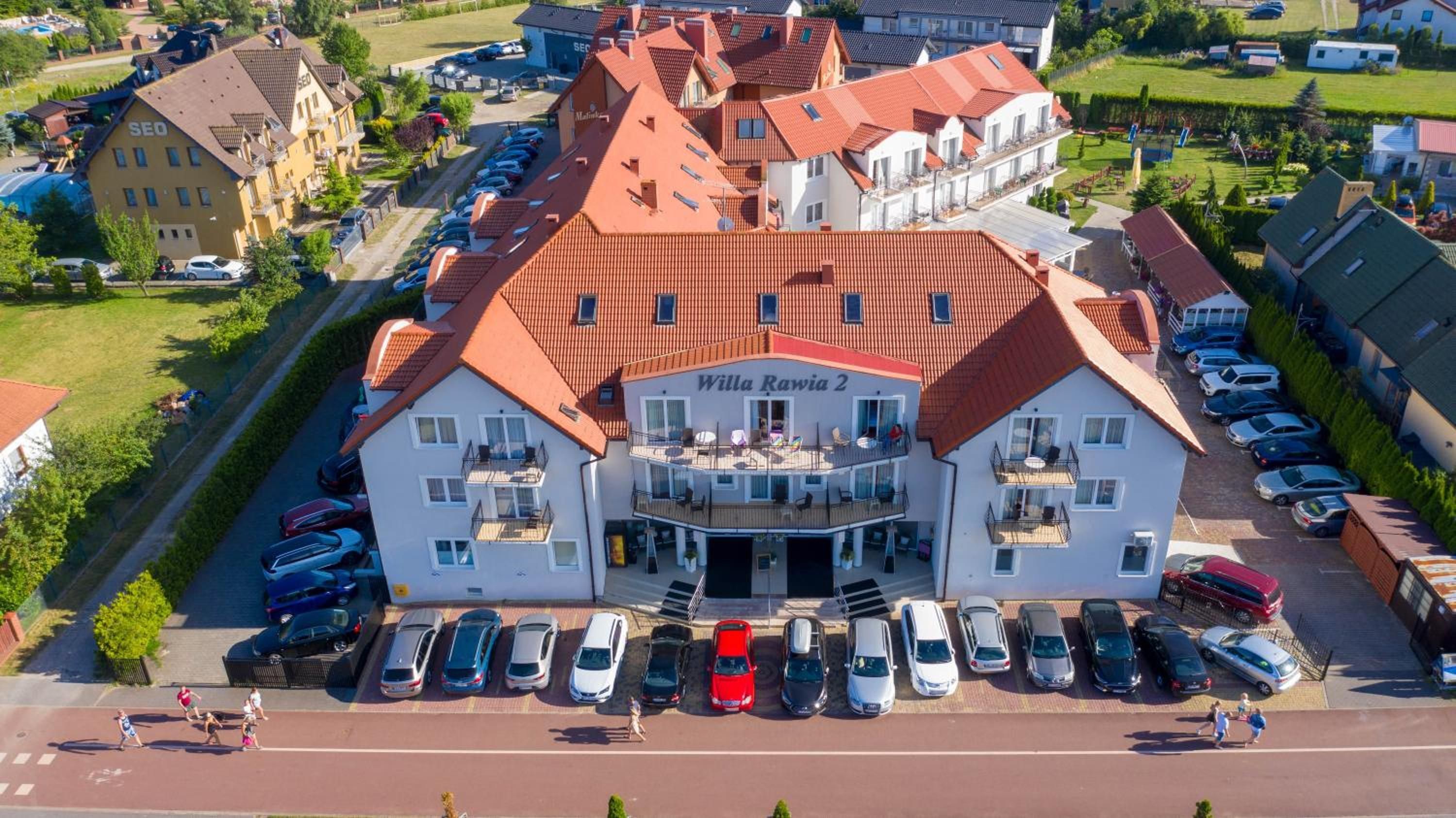 A two-story building with a red-tiled roof, labeled 'Willa Rawia 2,' featuring a central balcony and a parking lot with multiple cars.
