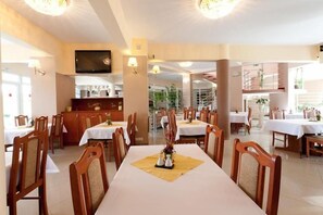 Un espace repas avec plusieurs tables et chaises, un téléviseur fixé au mur et, au fond, un coin cuisine.